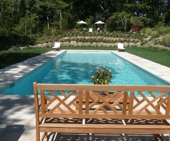 Wooden bench in front of a rectangular swimming pool surrounded by green grass, trees, and terraced landscaping.