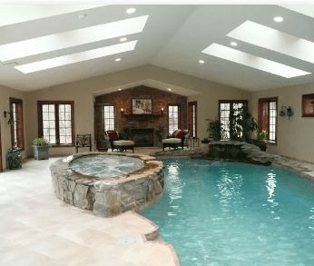 Indoor pool with hot tub, skylights, fireplace, and large windows in a beige-walled room.