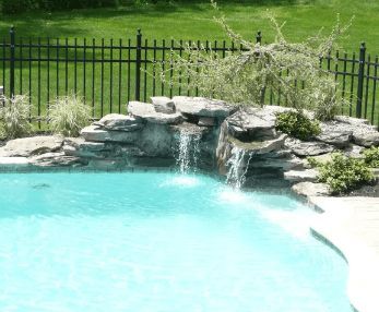 Pool with a rock waterfall feature, black fence, and green lawn.