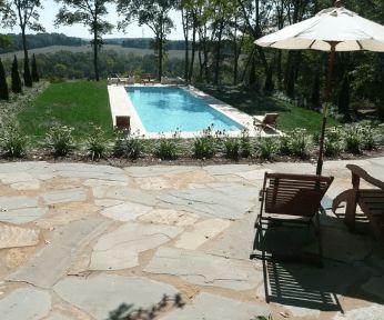 Stone patio with a pool, trees, and lounge chairs on a sunny day.