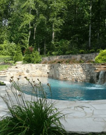 Swimming pool with waterfall and stone walls, surrounded by greenery and trees.