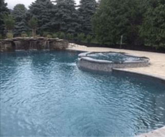 A pool with a built-in hot tub. A waterfall feature is visible in the background, trees surround the area.