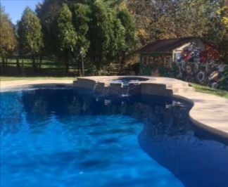 Blue swimming pool with a waterfall feature, surrounded by a concrete patio. A small building is in the background.