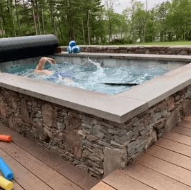 Person swimming in a small, rectangular spa with stone facing and wooden deck. Cover partially rolled up.
