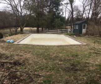 An empty rectangular pool with a beige surface surrounded by a brown border, in a grassy yard, with a shed.