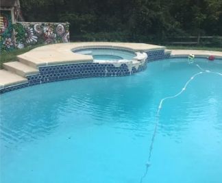 Swimming pool with a built-in hot tub, featuring blue tile and concrete steps; a garden backdrop.