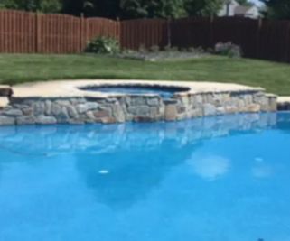 Blue pool with a stone-edged hot tub. Green grass, a brown wooden fence, and blue sky are in the background.
