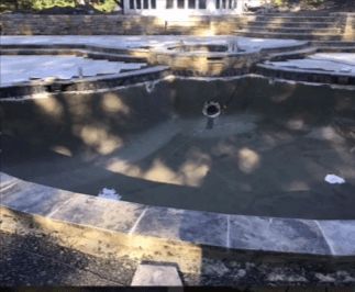 Empty, dark pool under construction, with stone edging and light reflections. White building in background.