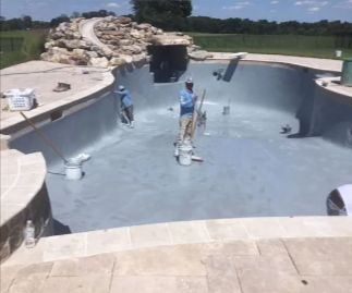 Two workers painting the inside of a gray, empty swimming pool. Landscaping and green field in background.