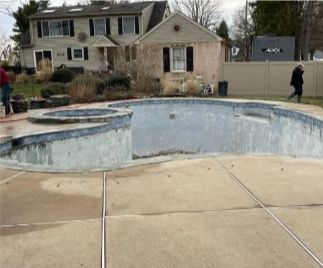 An empty, weathered swimming pool in front of a two-story house; a person walks beside a fence.
