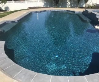 A dark-bottomed swimming pool with blue water surrounded by a grey stone border and a white fence.
