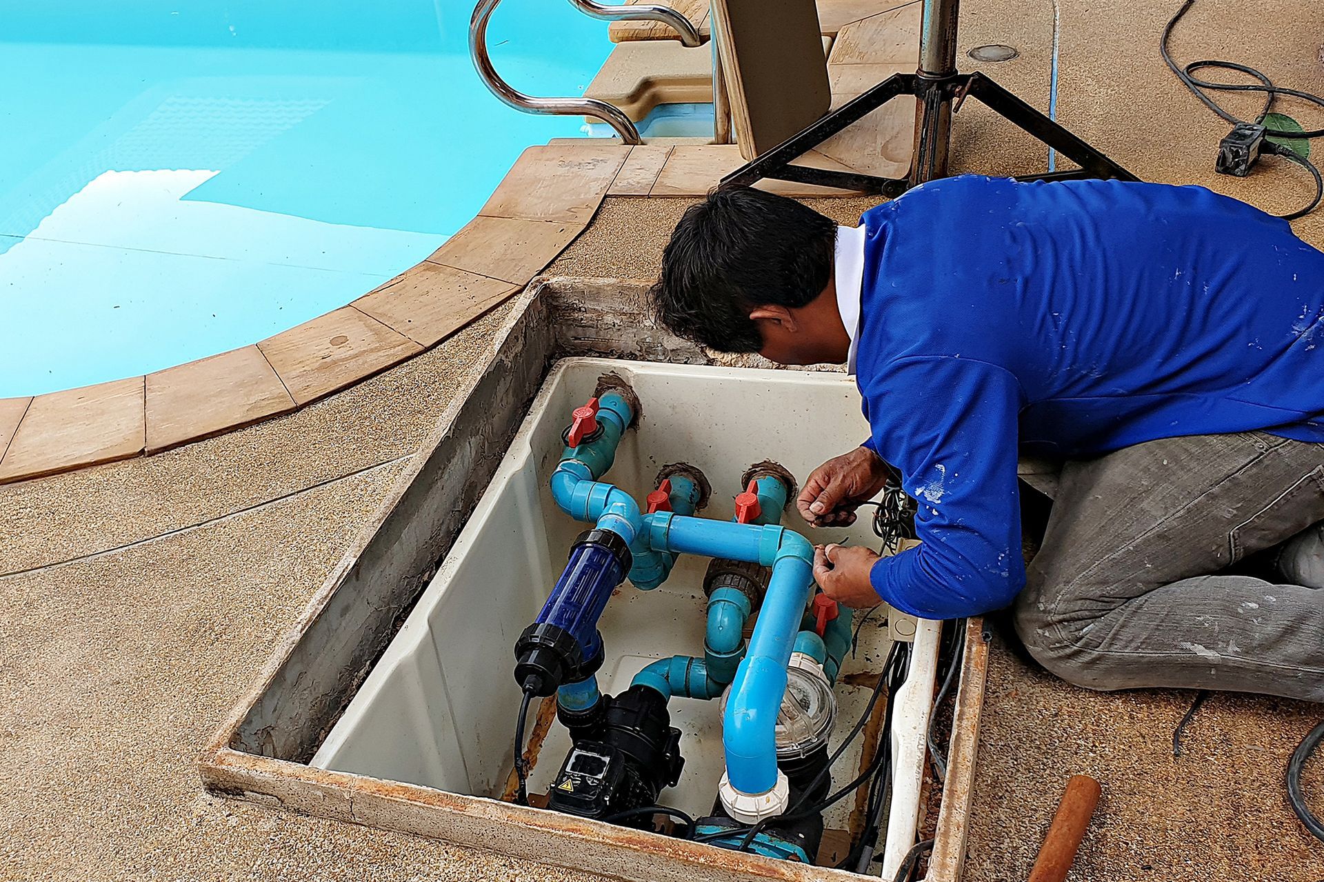 Man repairs pool equipment near pool, blue piping, open access hatch.