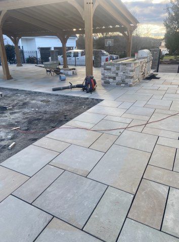 Paver patio under a wooden pergola during construction; a leaf blower is on the ground.
