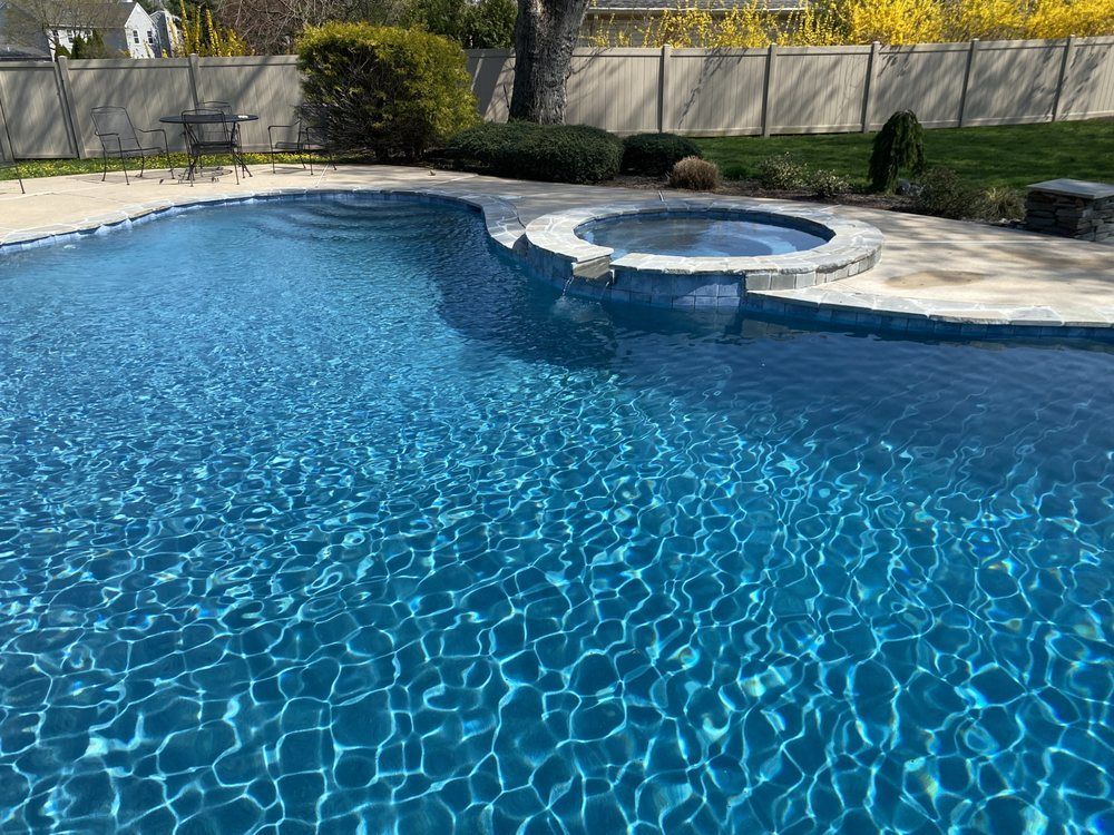 A shimmering blue swimming pool with a built-in hot tub.  Concrete deck with surrounding yard.