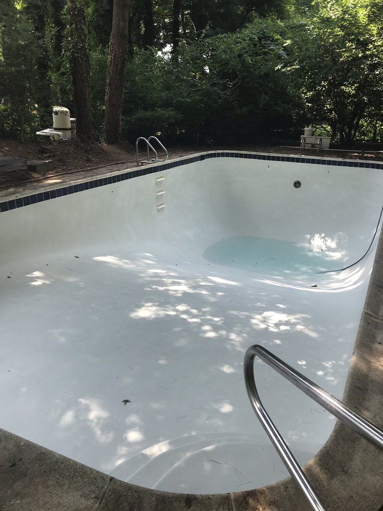 Empty, light-colored swimming pool with metal handrails and ladder, surrounded by concrete and greenery.