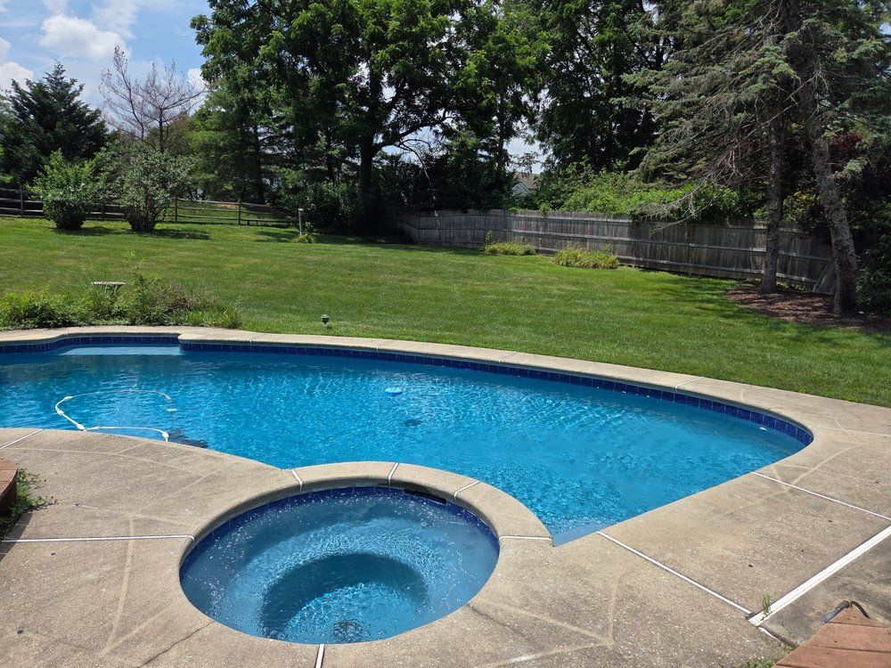 Backyard pool with a spa, surrounded by concrete. Green grass and trees in the background on a sunny day.