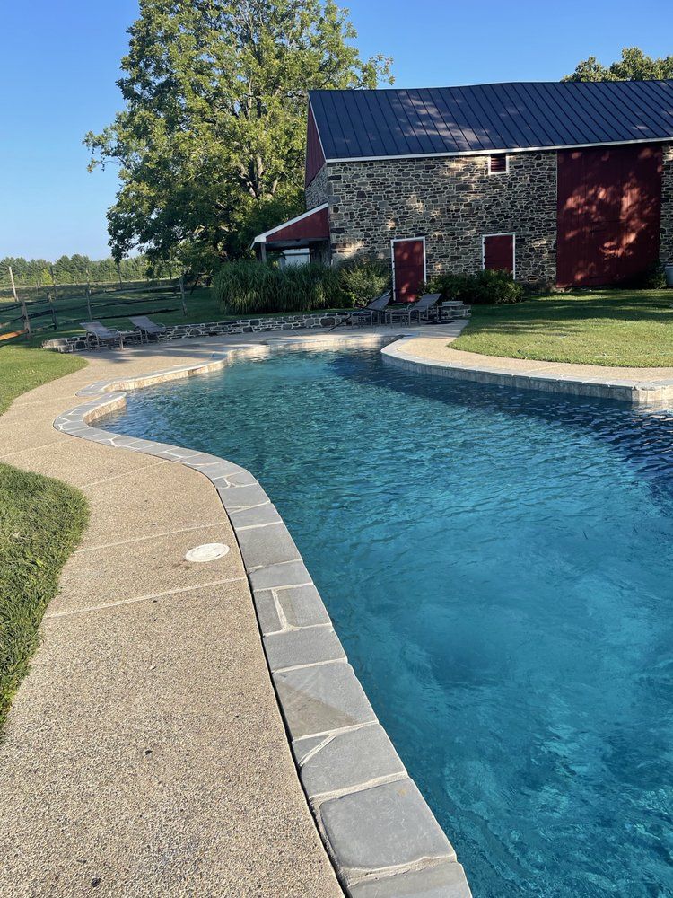 Swimming pool with blue water curves in front of a stone building with a red door and roof on a sunny day.