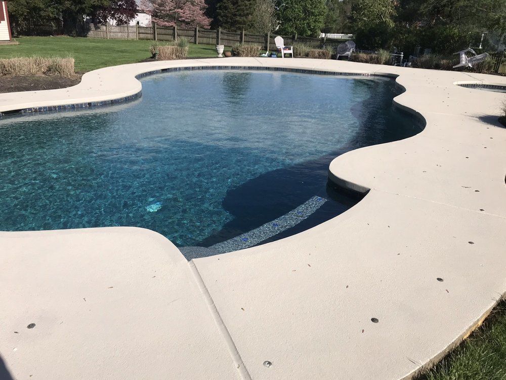 A freeform swimming pool with light concrete surround. Dark blue water reflects the sky.