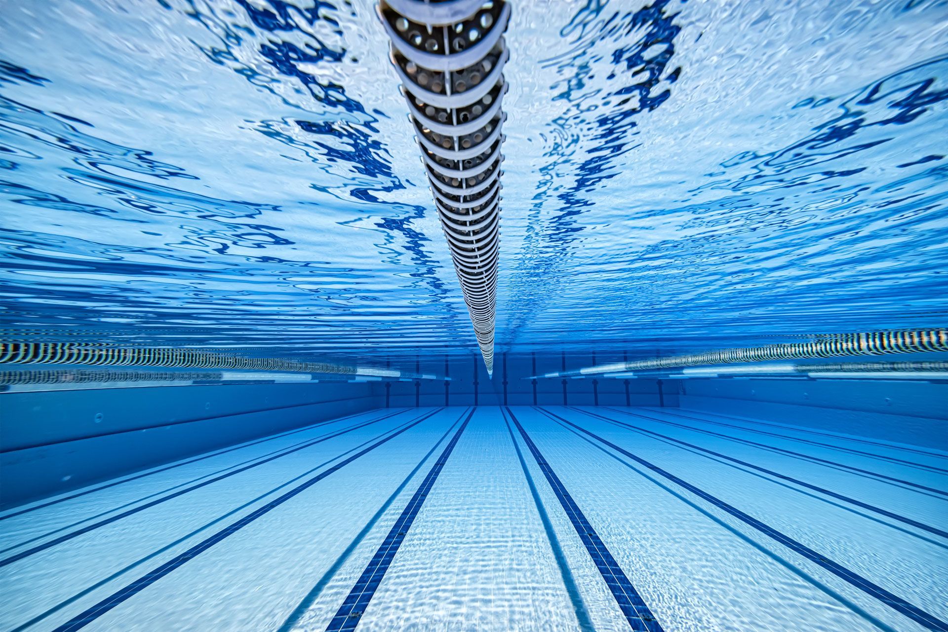 Underwater view of an empty swimming pool with lane markers and a central lane rope. Blue water and tiles.