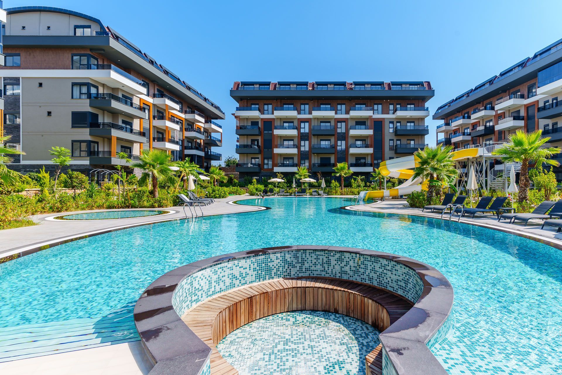 Swimming pool in front of apartment buildings with palm trees and a bright blue sky.
