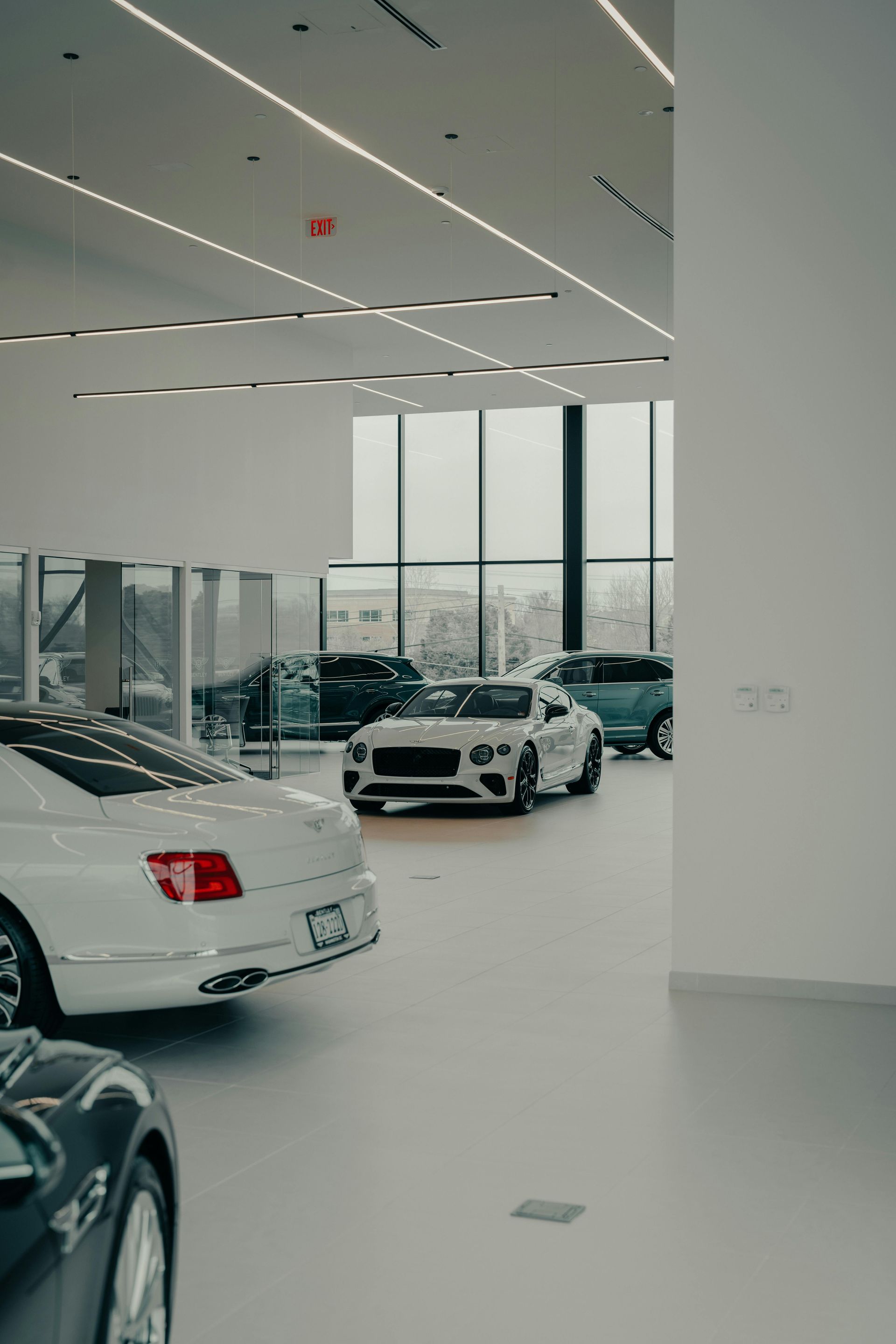 Luxury car showroom with white and dark vehicles, minimalist interior. Large windows, neutral tones.