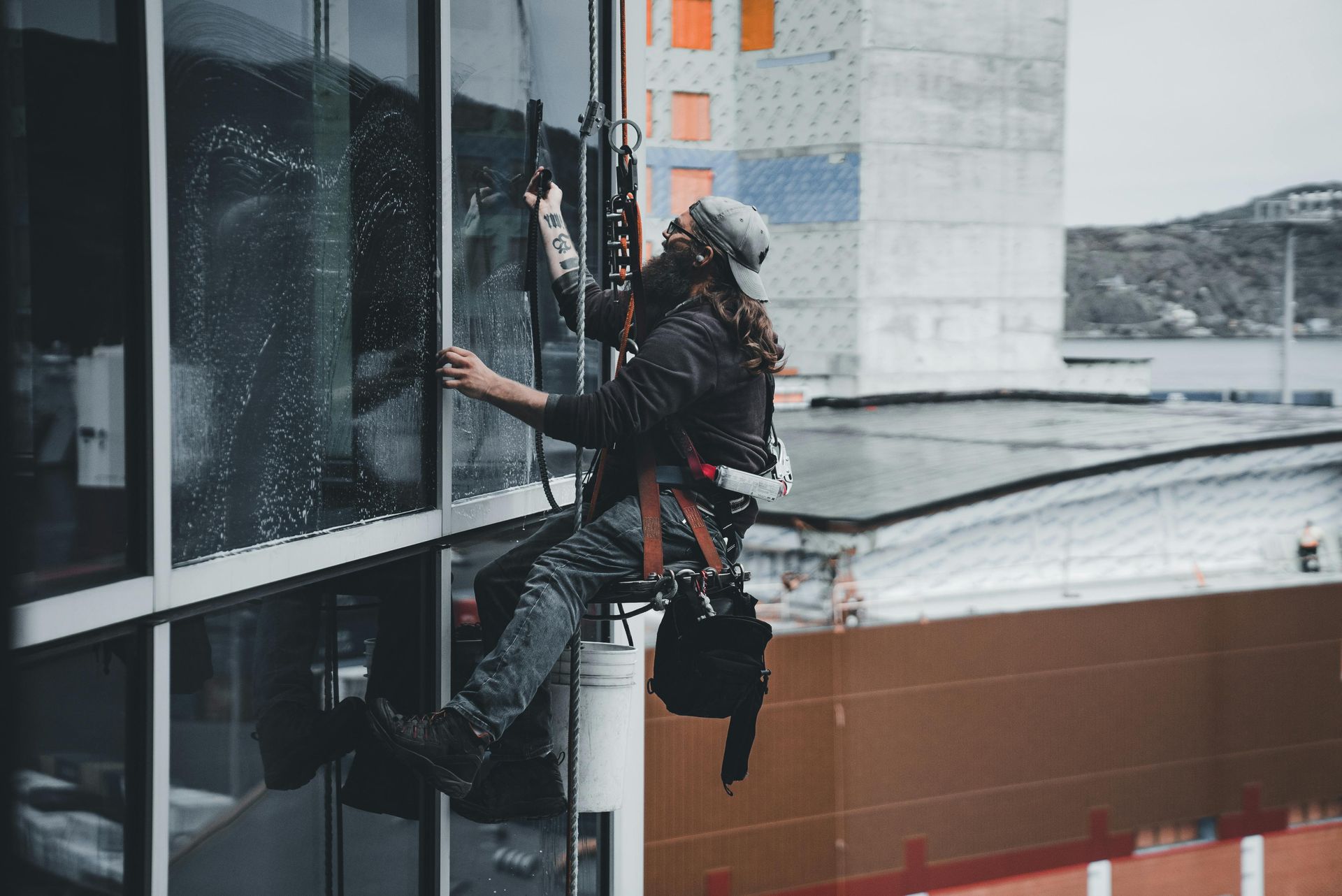 Person washing windows on a high-rise building, suspended by ropes, wearing safety gear.