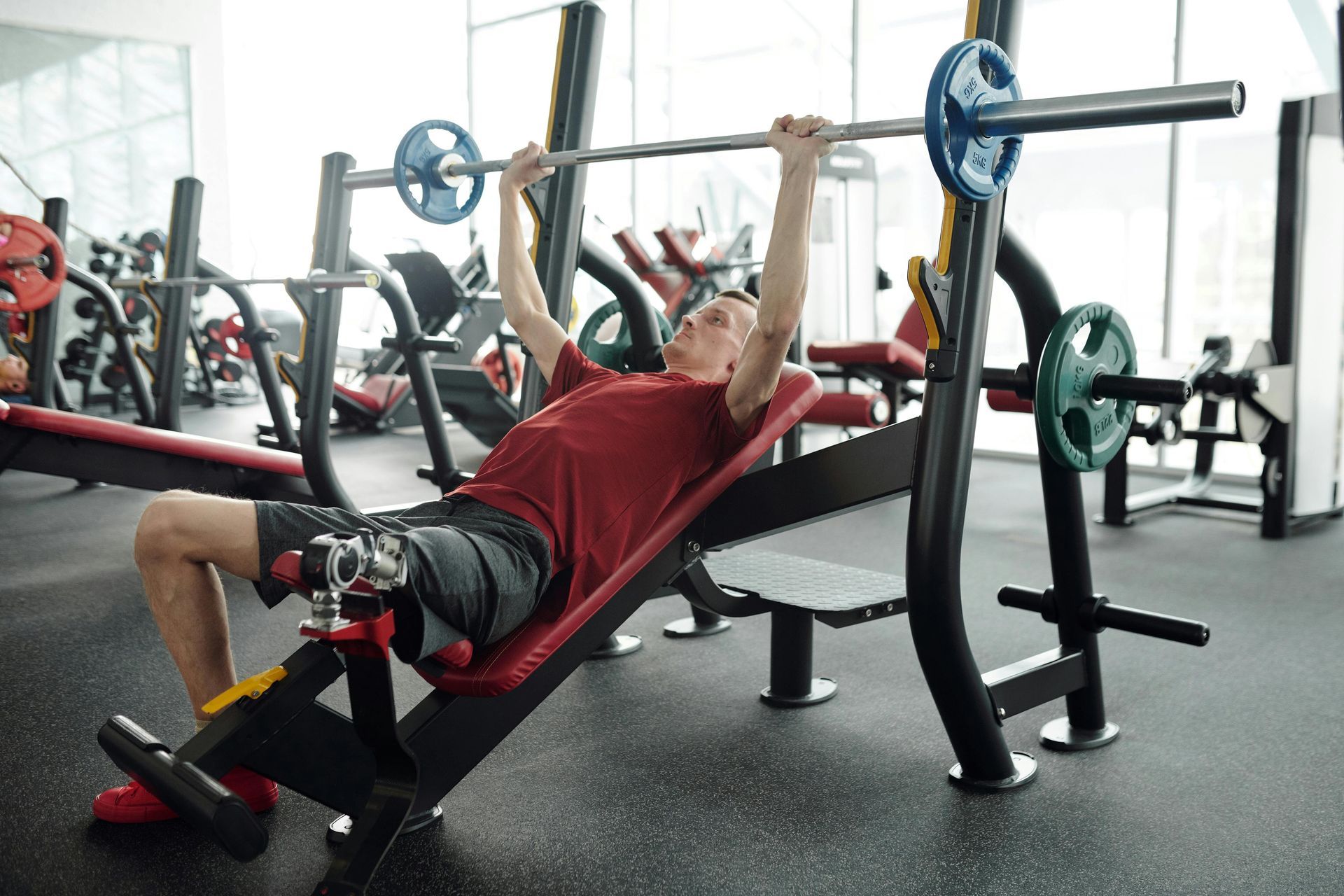 Man in red shirt bench presses with barbell in gym.