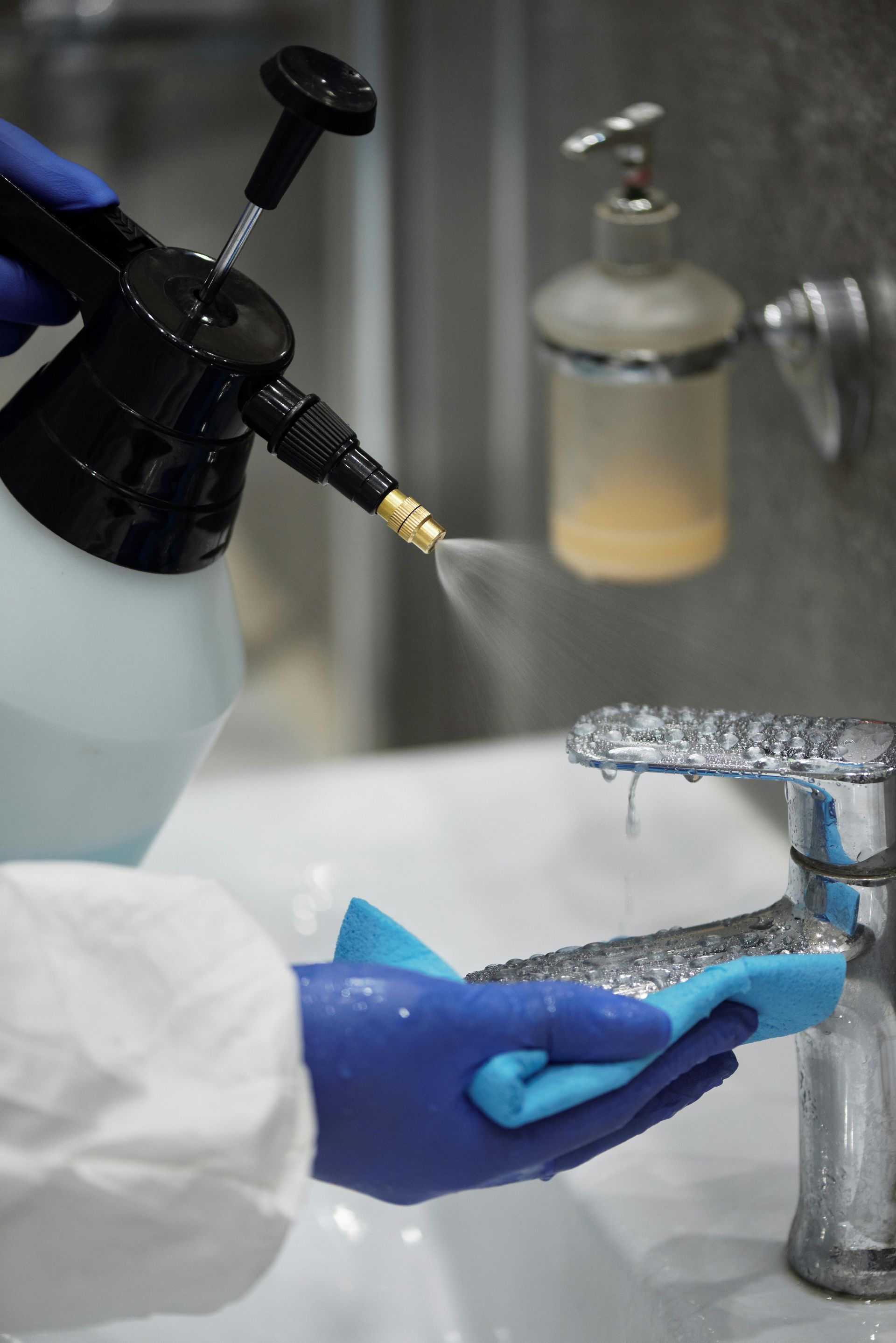 Person in blue gloves spraying and wiping a chrome faucet in a bathroom.