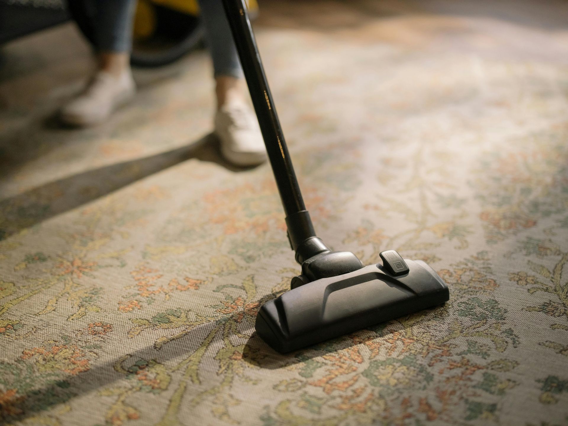 Vacuum cleaner cleaning a patterned carpet, with a person's legs and white shoes in the background.