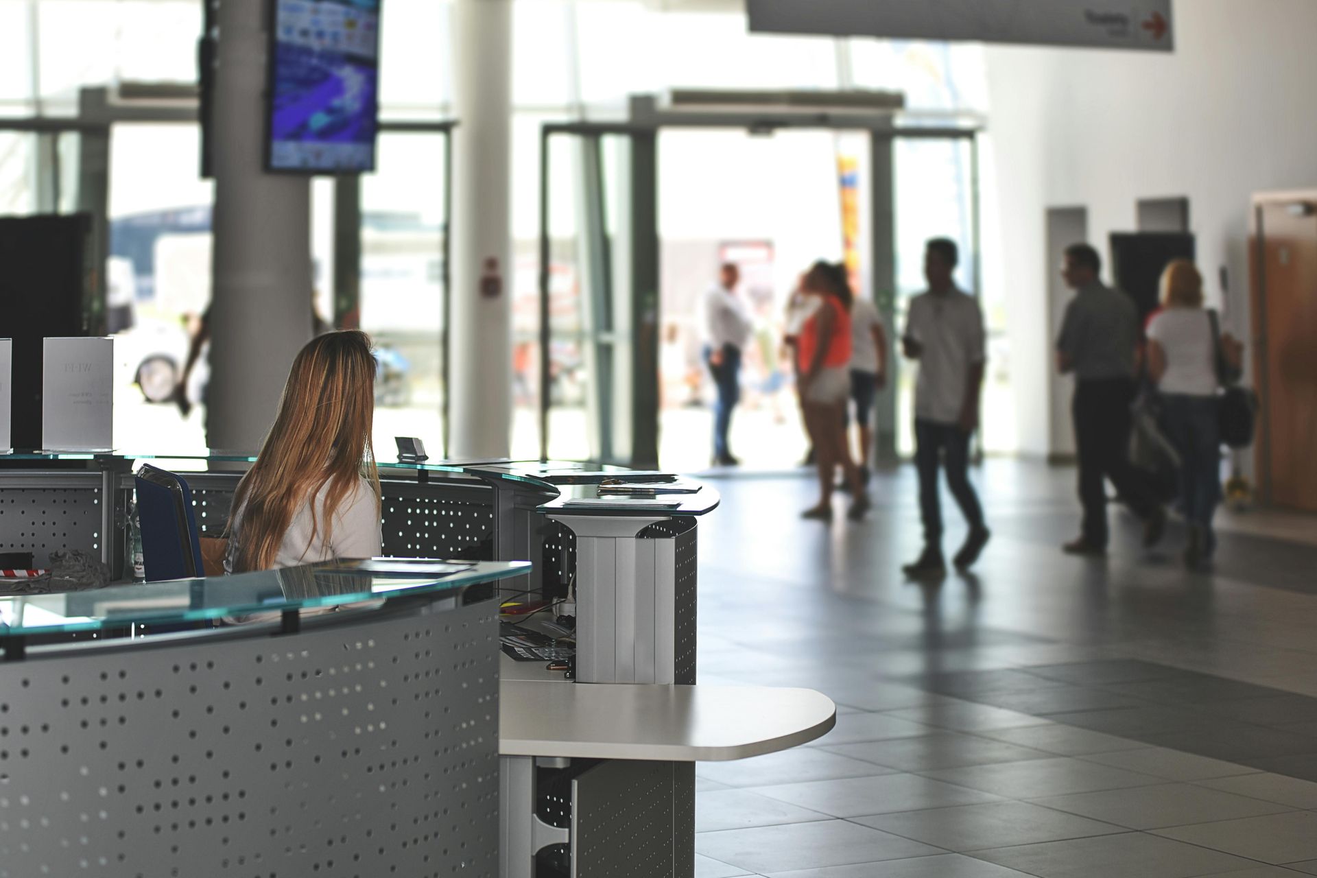 Woman at reception desk in a modern building, with blurred figures entering and exiting through glass doors.