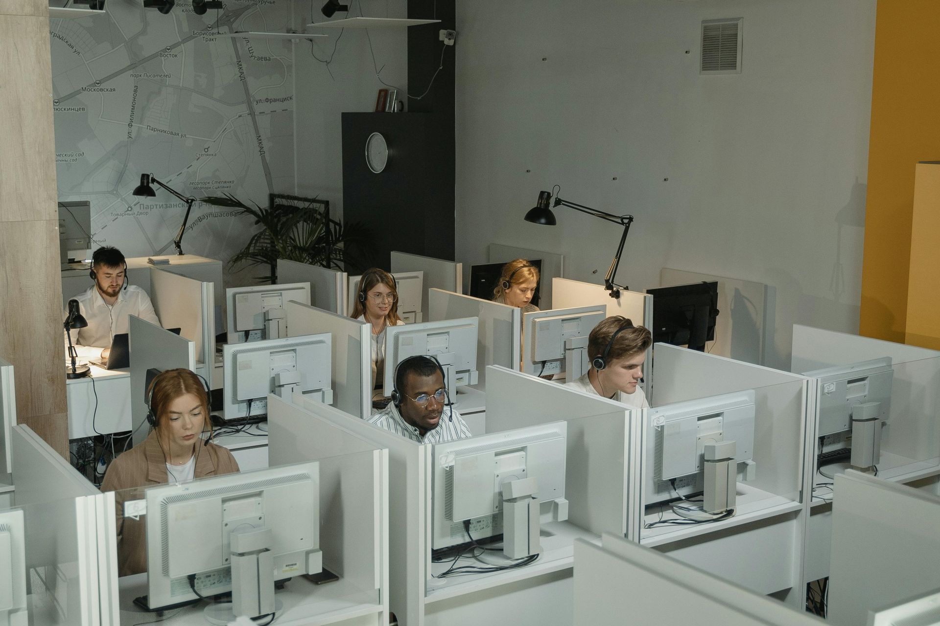 People working at computers in an office with cubicles; natural light and overhead lamps.