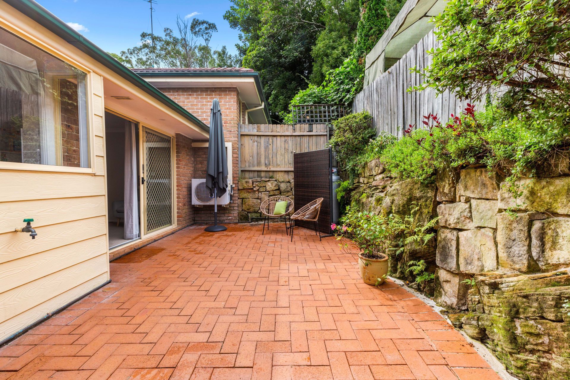 A brick patio with a chair and umbrella in front of a house.