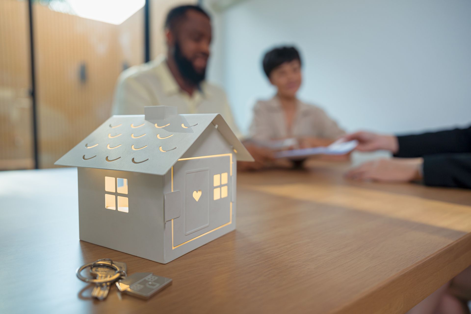 Small house model with keys on table, people reviewing documents behind. Small house model with keys on table, people reviewing documents behind.