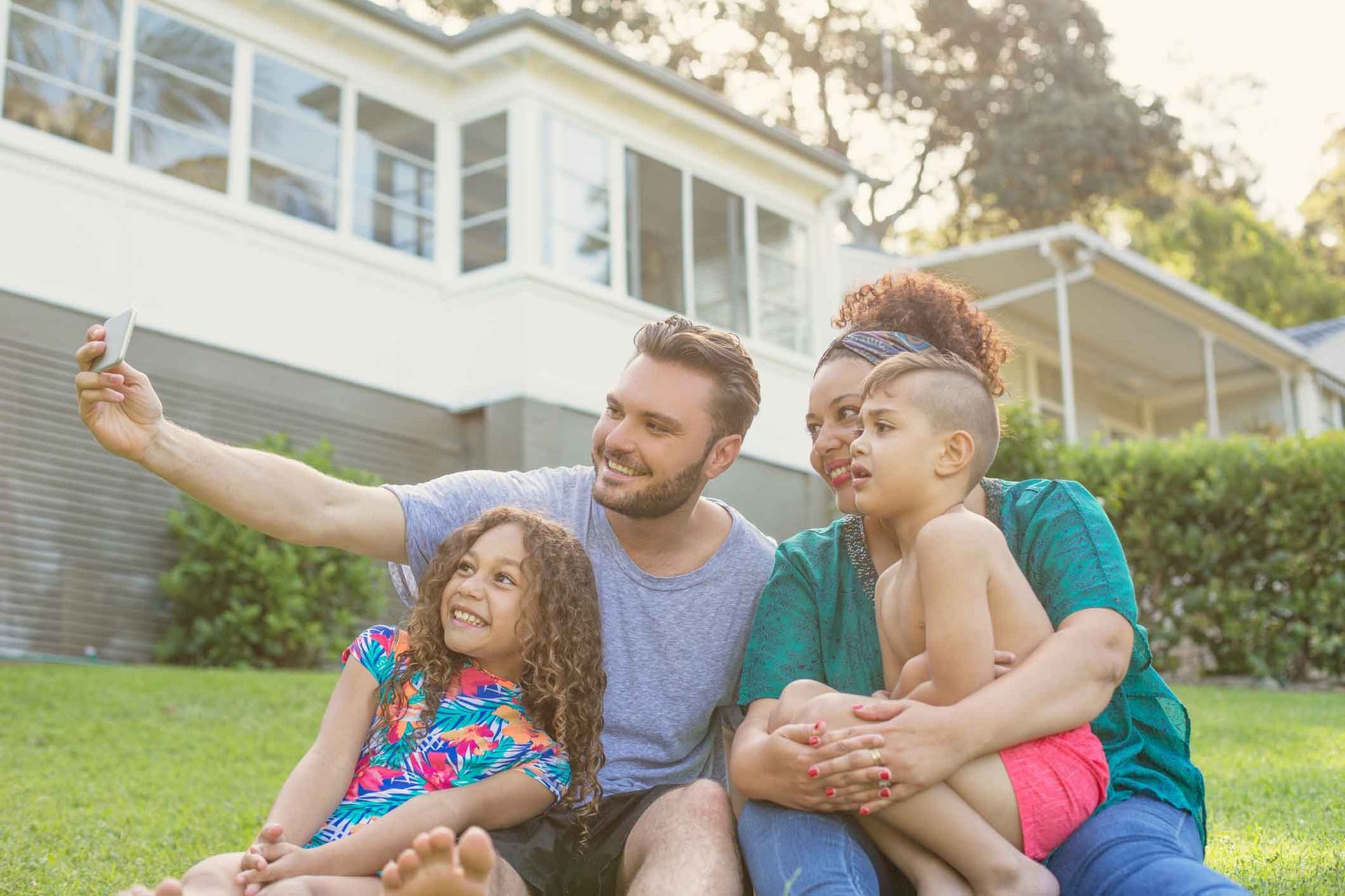 A family is sitting on the grass in front of their house taking a selfie.