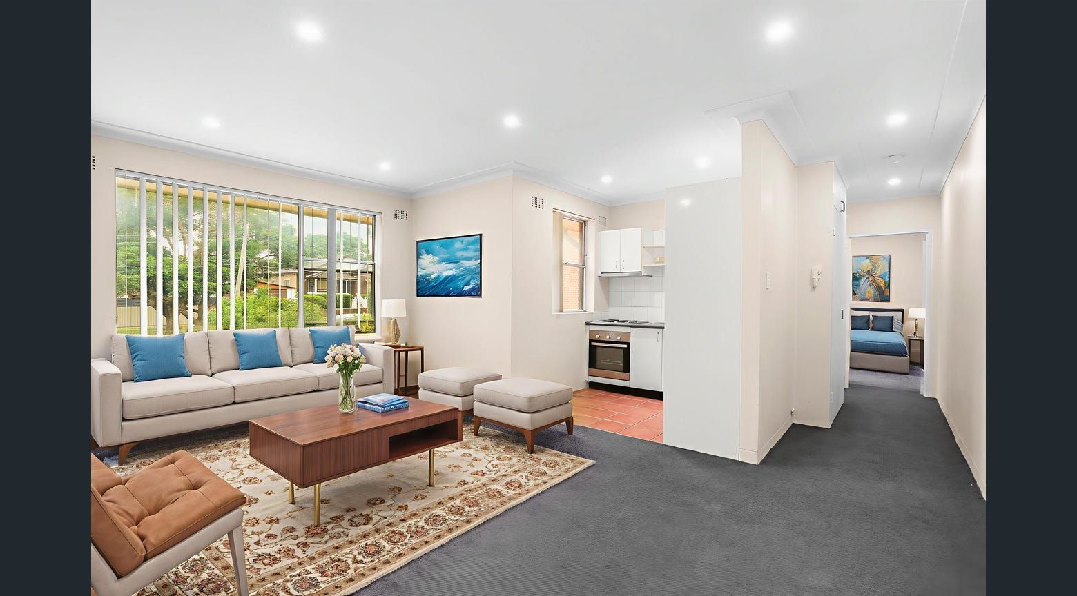 Living room with sofa, coffee table, and open kitchen. Neutral tones with a blue accent painting.