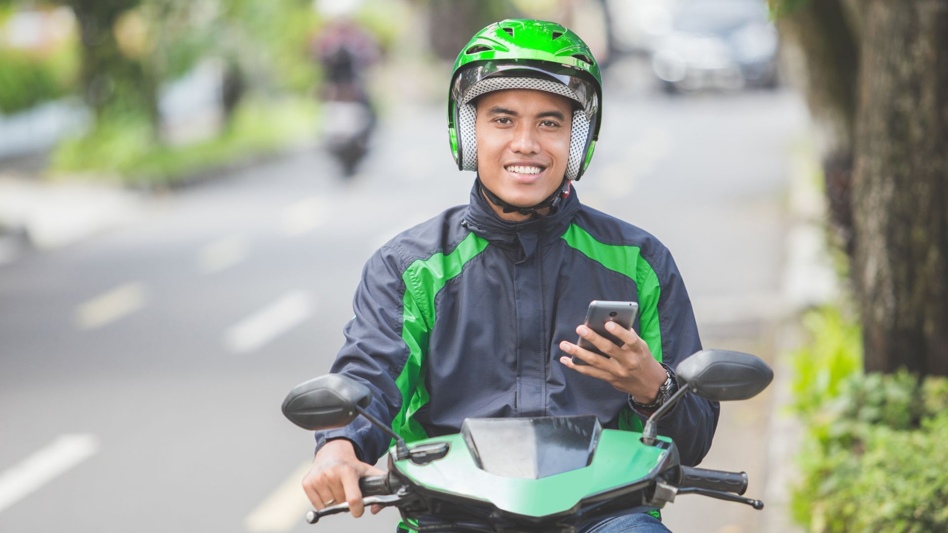 Smiling delivery rider in green helmet and jacket on a scooter, holding a phone on a city street
