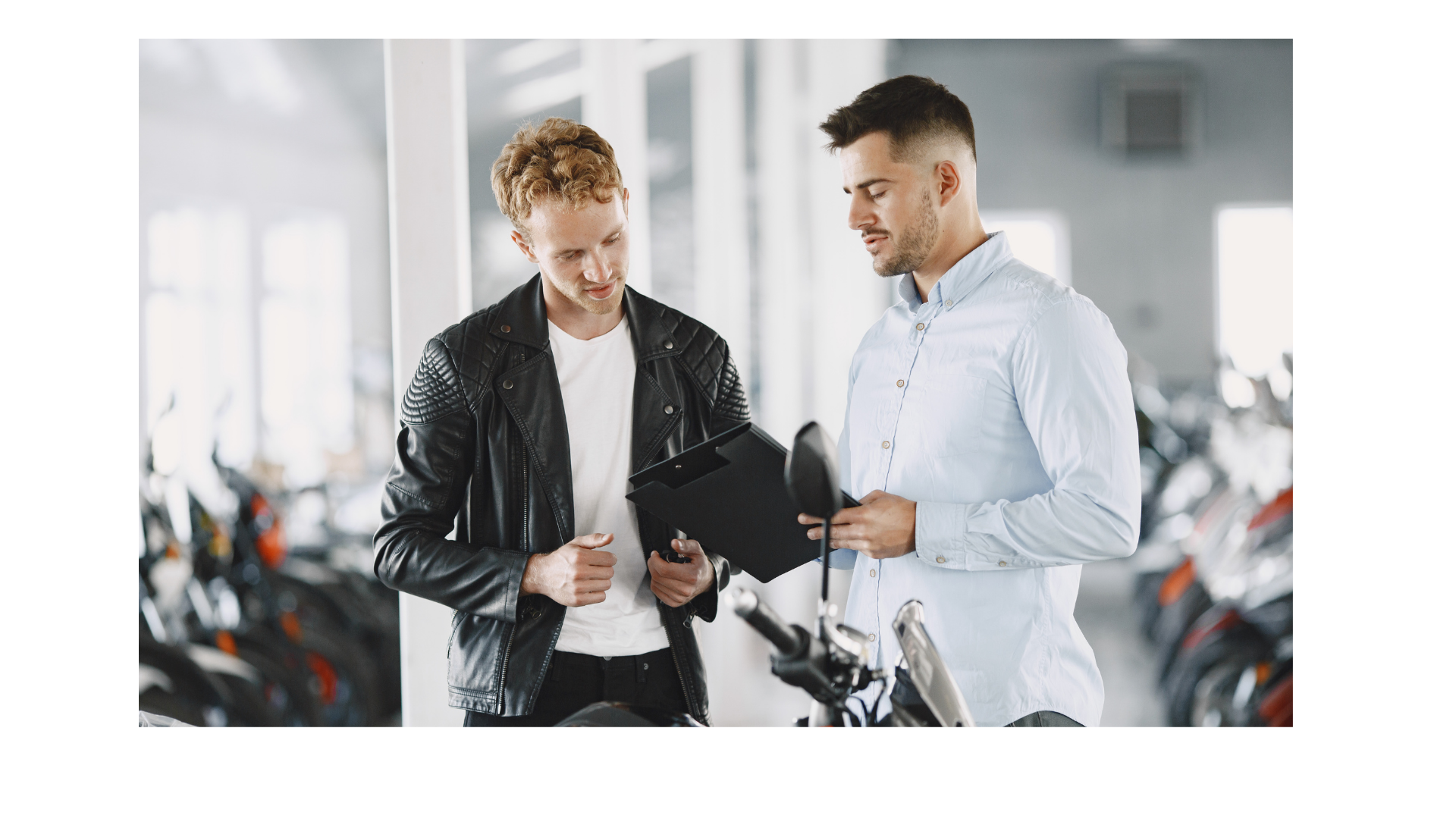 Two men discussing a tablet in a motorcycle showroom, with bikes visible in the background.