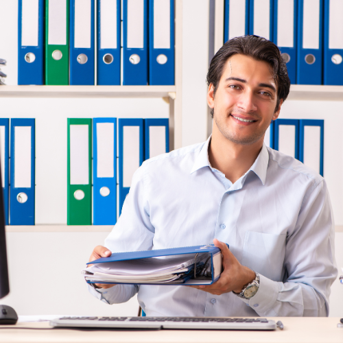 A man is sitting at a desk in front of a computer holding binders.
