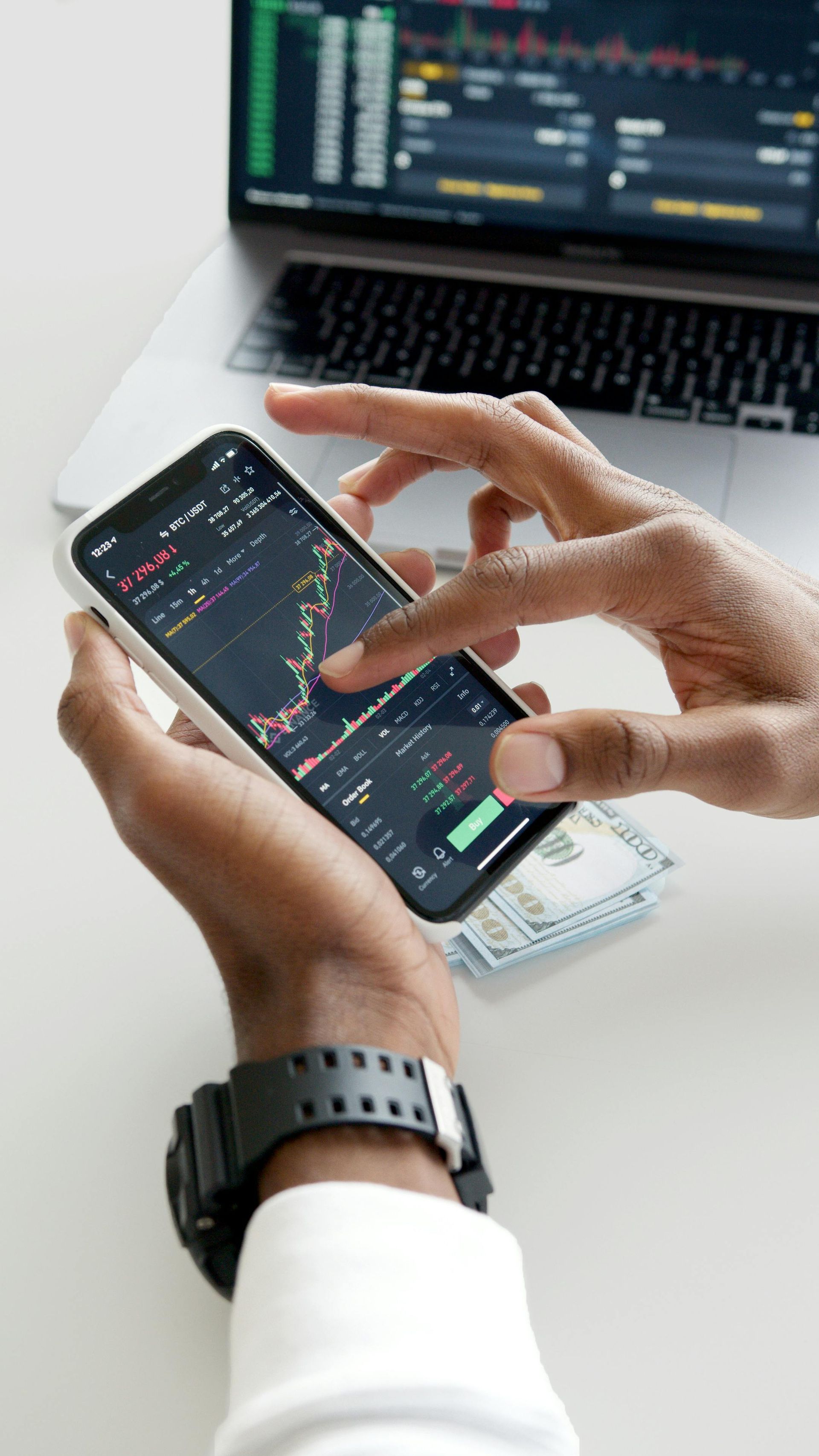 Person using a smartphone to analyze stock charts, with a laptop and cash visible.