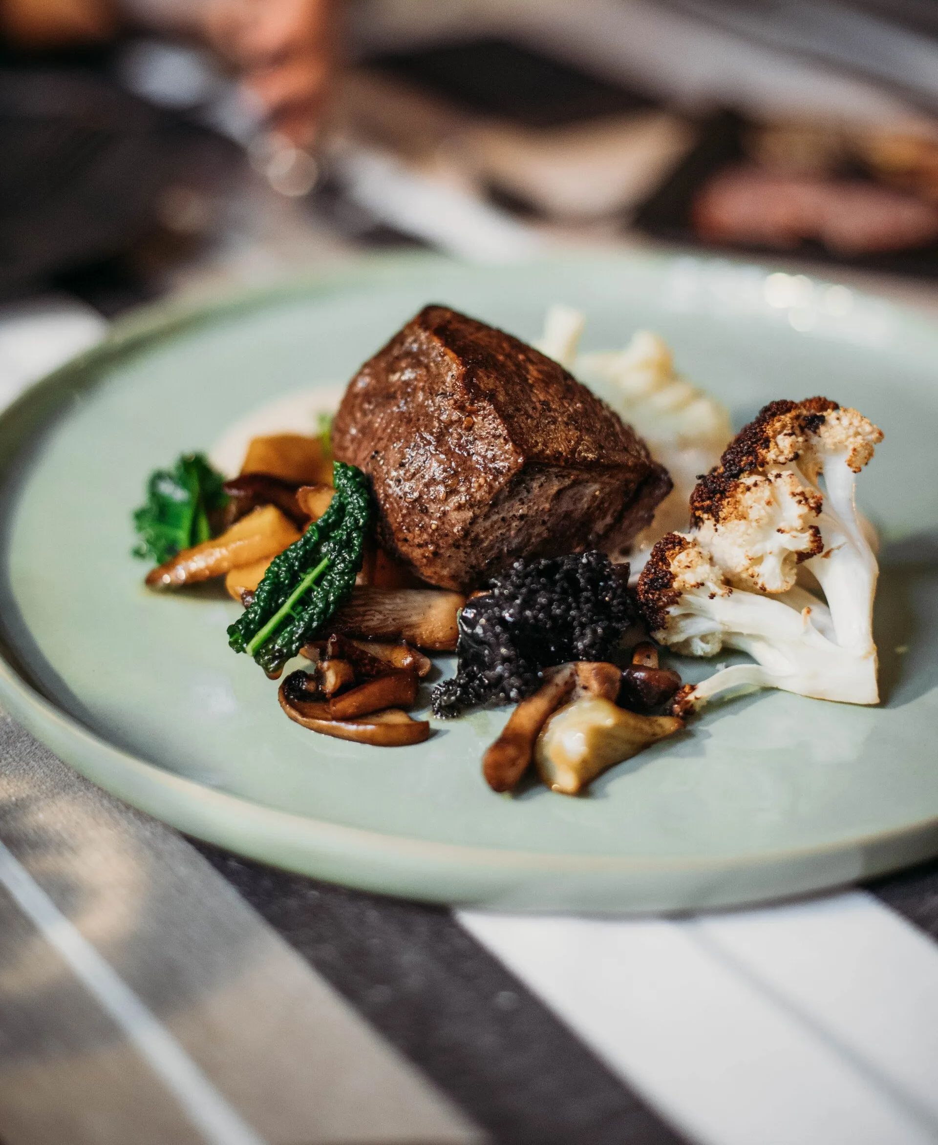 Plate of seared steak, cauliflower, mushrooms, and greens on a light green plate, striped tablecloth.