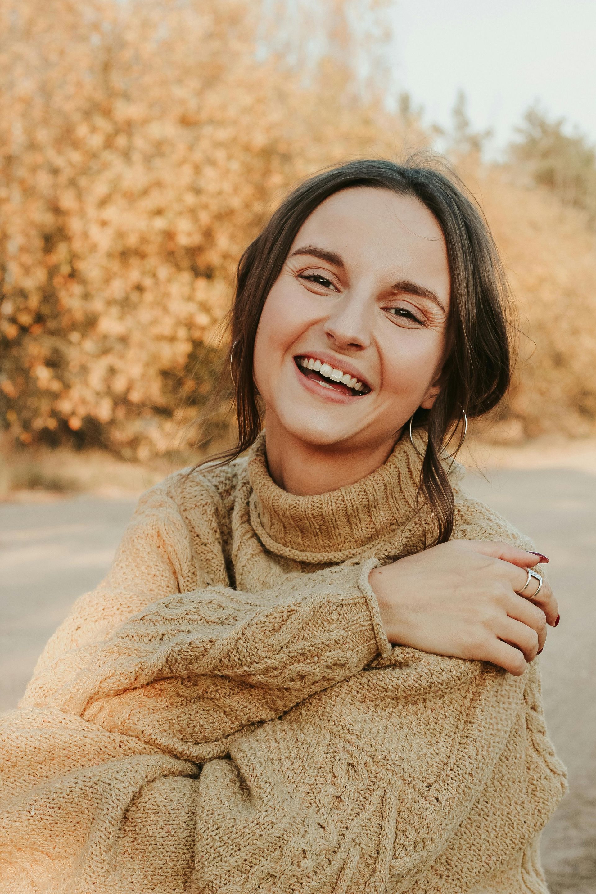 Woman in beige sweater smiles outdoors, fall foliage in background.