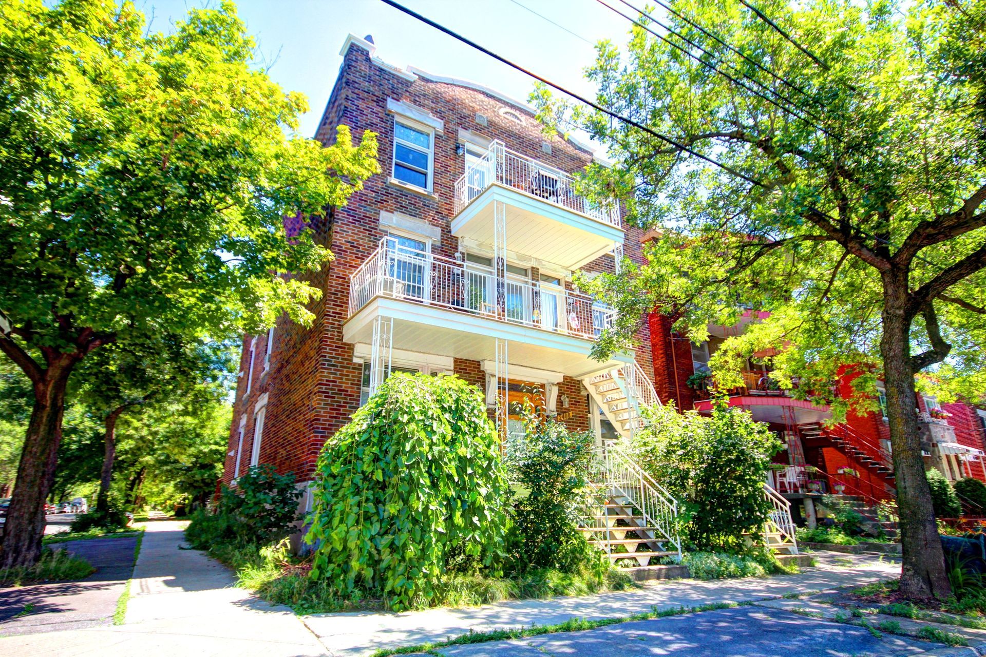 Un grand bâtiment en brique avec balcon est entouré d'arbres par une journée ensoleillée.