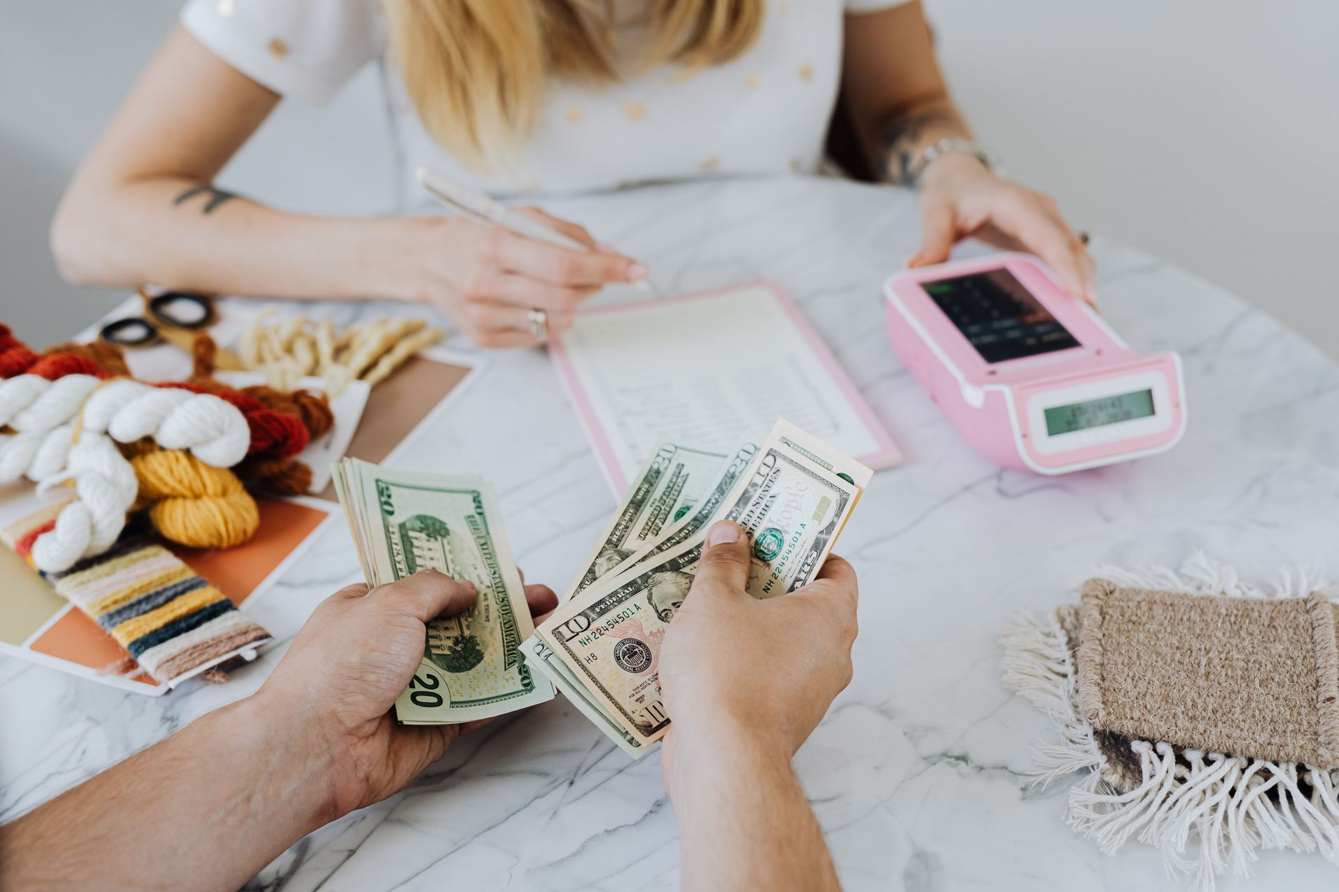 Hands counting cash at a tabletop with a pink card reader, crafts, and a notepad on a marble surface