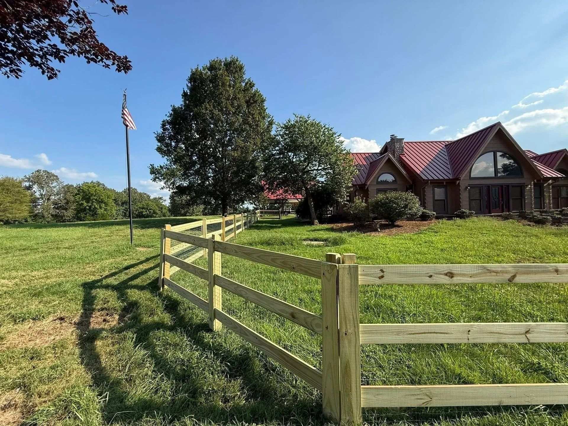 Wooden fence in a grassy field with a house and an American flag in the background.