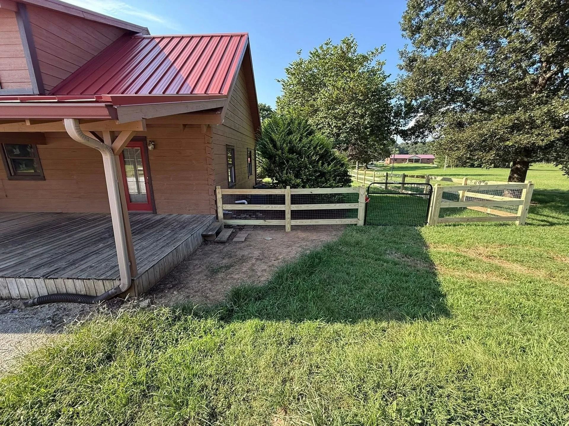 Tan house with red roof, wooden deck, and a new split-rail fence enclosing grass area. Sunny day.