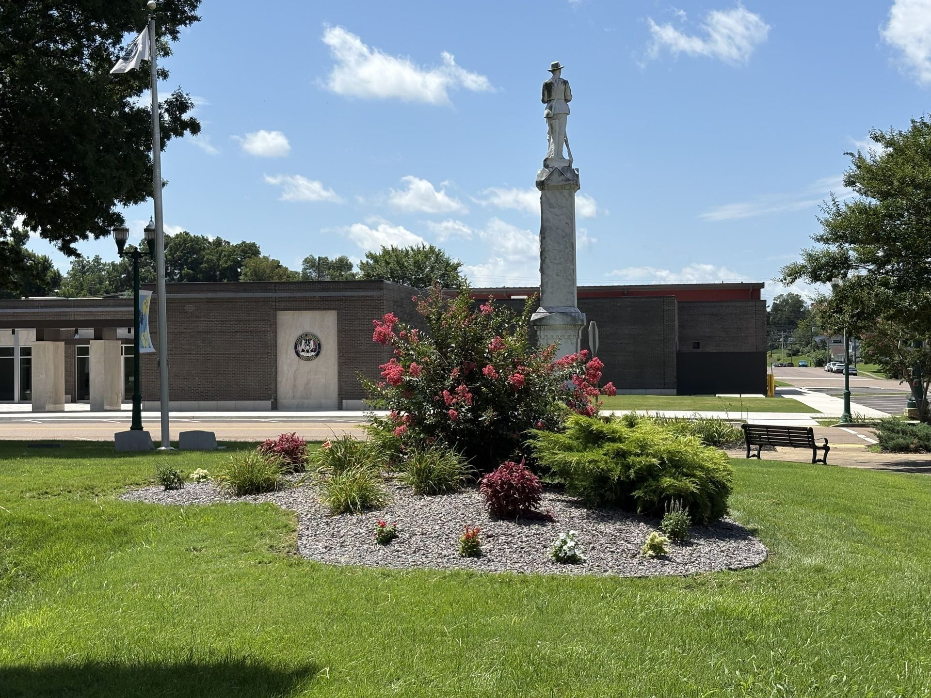 Park view with a flower bed in front of a brick building and a tall monument under a blue sky.