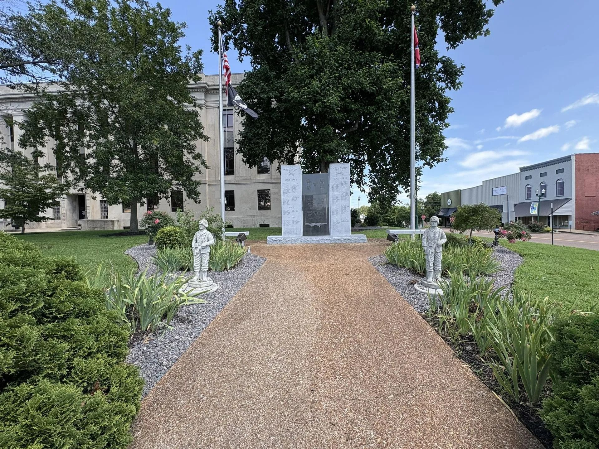 A gravel path leads to a war memorial flanked by statues and flags, with a courthouse and buildings in the background.