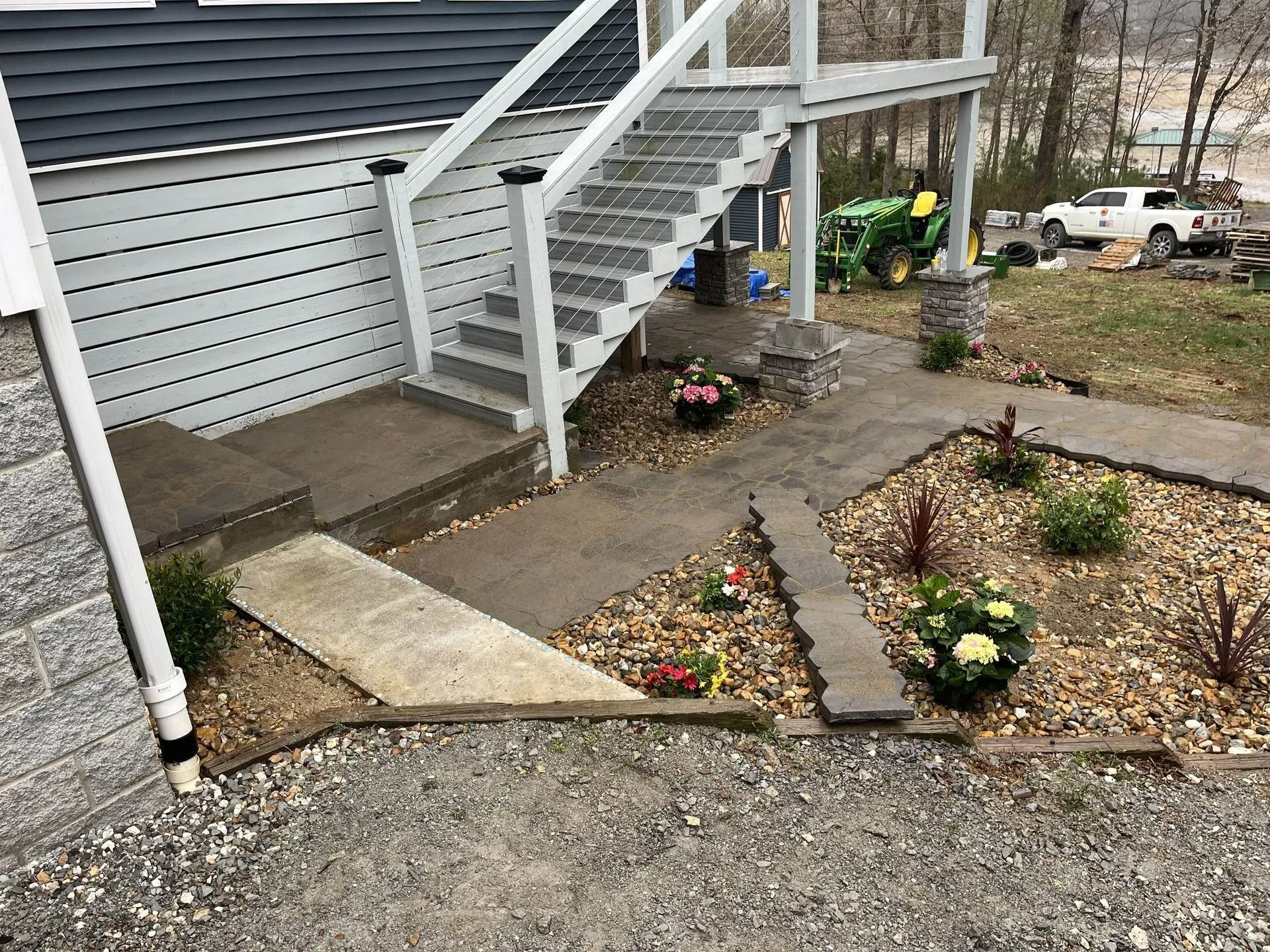 Concrete steps and walkway leading to a house with a raised deck, gravel ground, and landscaping.
