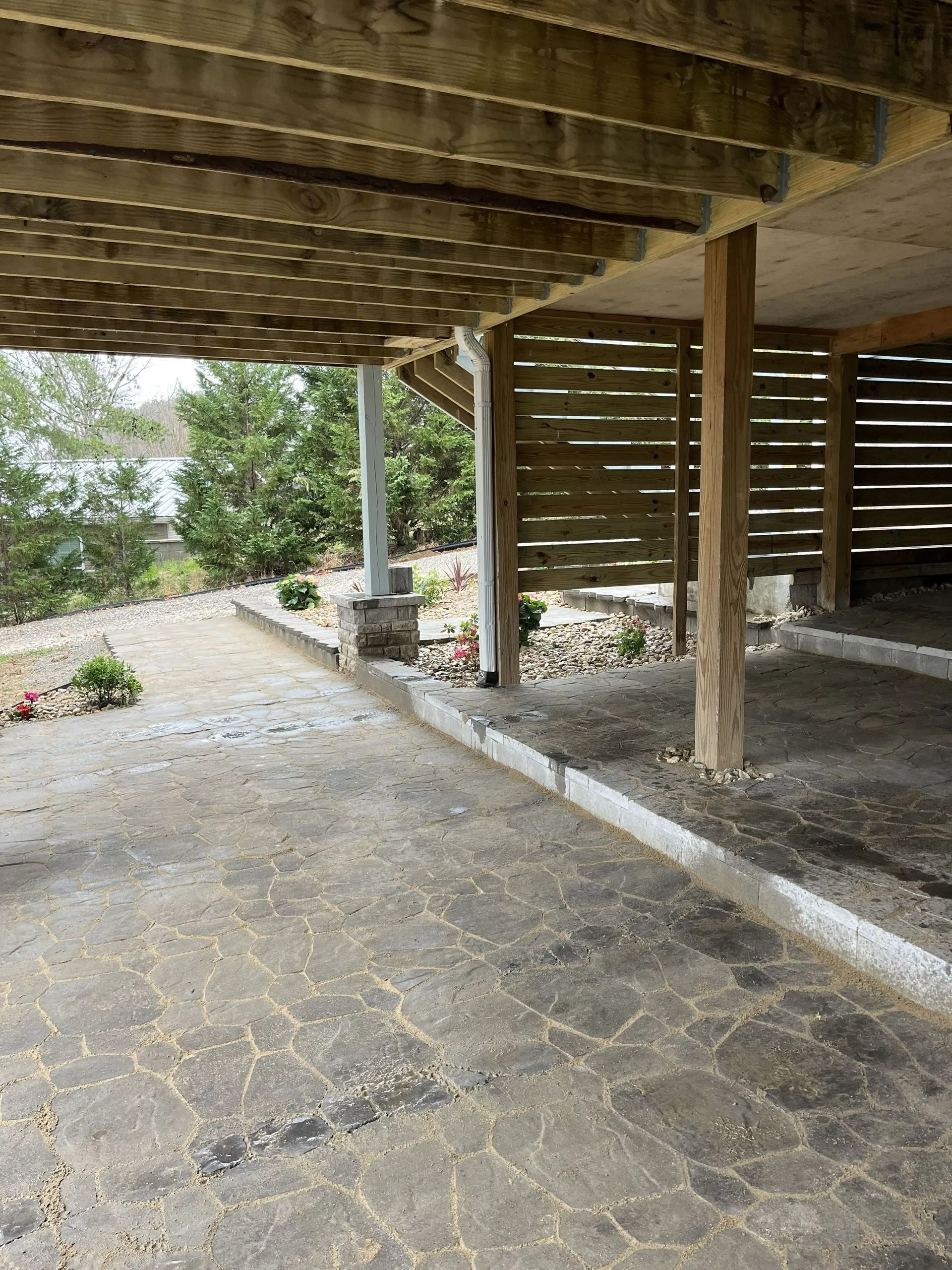Underneath a wooden deck, a concrete path leads to a gravel area near a body of water.