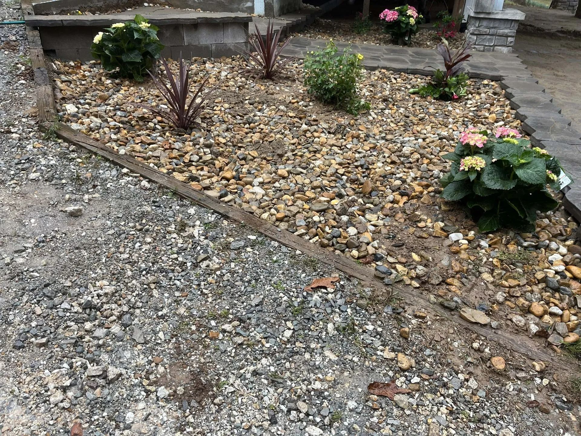 A rock garden bed with plants, including pink and yellow flowers, and red foliage, next to a gravel path.